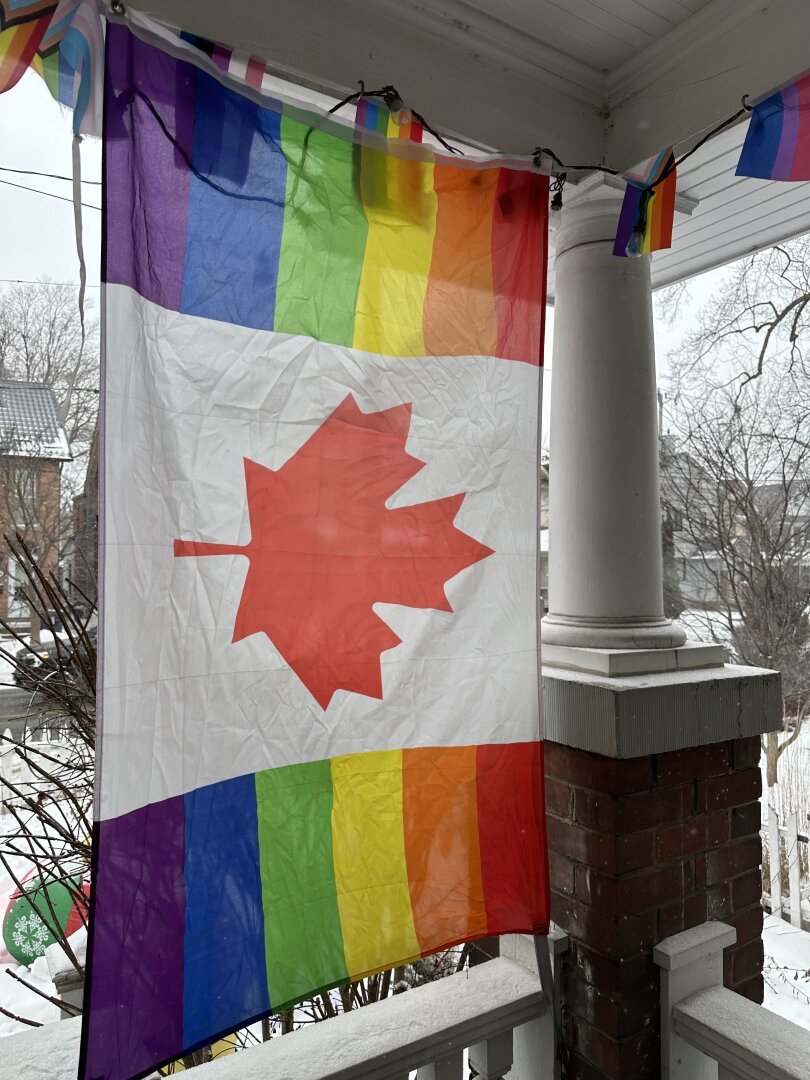 A rainbow Canadian flag hanging on a porch. The neighbourhood behind is snowy.