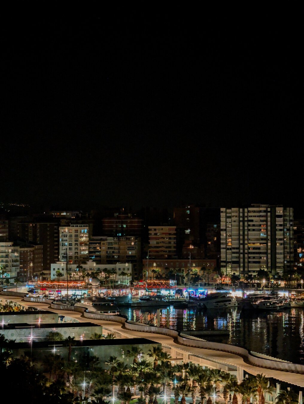 Night view of the Malaga's marina harbour, there is a strong contrast between th light of the city buildings and the dark of the background.