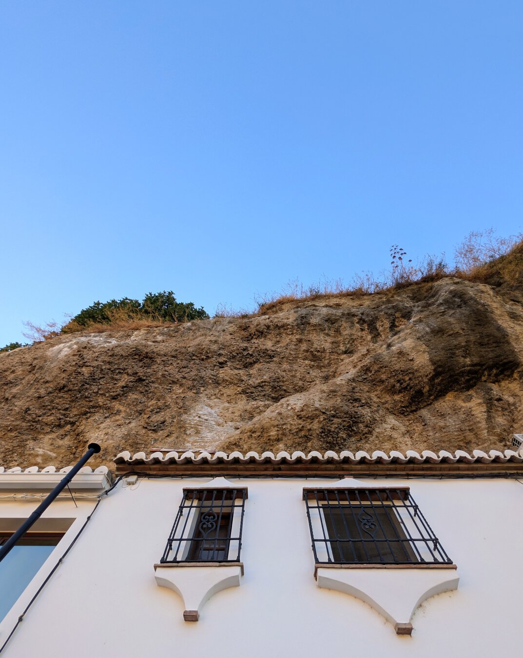 Contrast between the white wall of a house, the grey colour of the rocks above it and the bluse sky above the rocks.