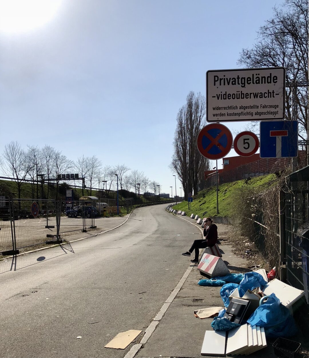 Das Bild zeigt eine asphaltierte Straße, die sich leicht nach links biegt. Am rechten Straßenrand sitzt eine Person auf einem Betonblock. Vor ihr liegt Müll in blauen Säcken und Kartons. Im Hintergrund stehen mehrere Verkehrsschilder, darunter ein Schild mit der Aufschrift "Privatgelände - videoüberwacht - widerrechtlich abgestellte Fahrzeuge werden kostenpflichtig abgeschleppt". Links von der Straße verläuft ein Zaun, dahinter befindet sich ein Gelände mit geparkten Fahrzeugen und Bäumen. Der Himmel ist hellblau mit leichten Wolken.

Bereitgestellt von @altbot@fuzzies.wtf, generiert mit Gemini