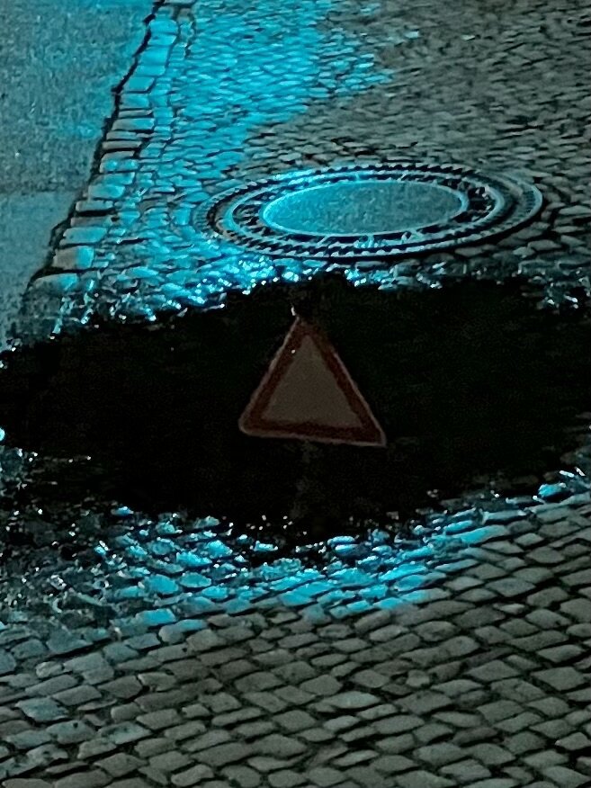 Das Bild zeigt eine dunkle Szene auf einem gepflasterten Weg. Ein Gullydeckel ist zu sehen, umgeben von einer dunklen, vermutlich nassen Fläche, die wie eine tiefe Pfütze wirkt. In der Mitte dieser dunklen Fläche ist das Spiegelbild eines dreieckigen Warnschildes erkennbar, das vermutlich in der Nähe steht. Das Pflaster und der Gullydeckel sind durch ein schwaches, blaues Licht beleuchtet, was der Szene eine unheimliche Atmosphäre verleiht.

Bereitgestellt von @altbot, generiert mit Gemini