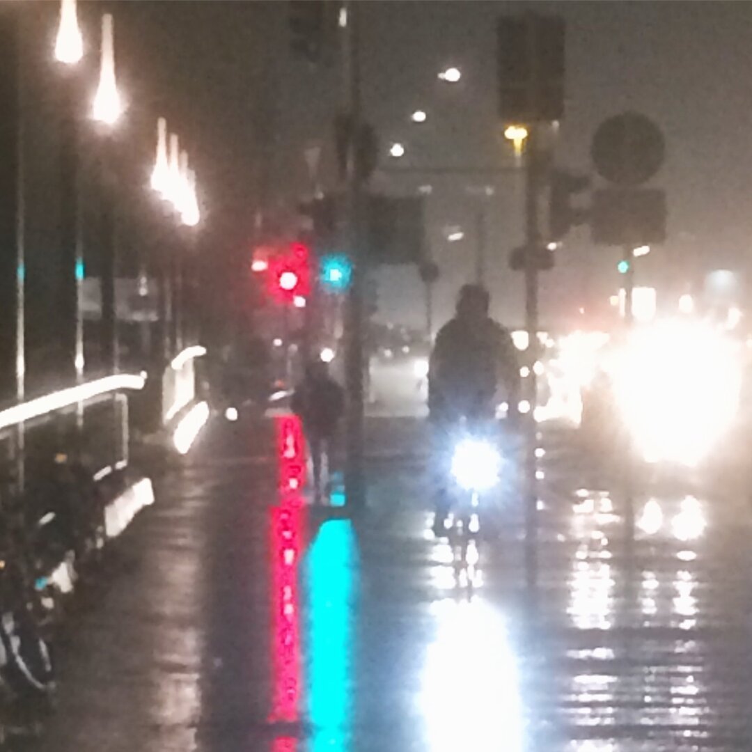 Das Bild zeigt eine nächtliche Straßenszene bei Regen. Die Oberfläche ist nass und reflektiert das Licht der Straßenlaternen und Ampeln. Im Vordergrund fährt eine Person mit dem Fahrrad, dessen Scheinwerfer hell leuchten. Im Hintergrund sind weitere Fahrzeuge und Fußgänger zu sehen, sowie Ampeln, die rot und grün leuchten. Am linken Bildrand befindet sich ein Geländer, an dem Fahrräder abgestellt sind. Die Szene wirkt durch den Regen und das Licht etwas verschwommen.

Bereitgestellt von @altbot, generiert mit Gemini