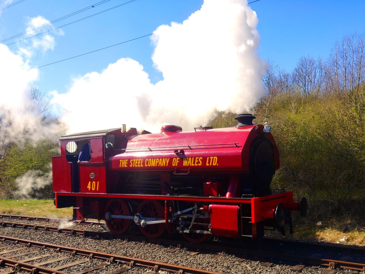 A steam locomotive on a sunny day. The engine is red and has The Steel Company of Wales Ltd. in yellow lettering. There is lots of white steam against a blue sky.