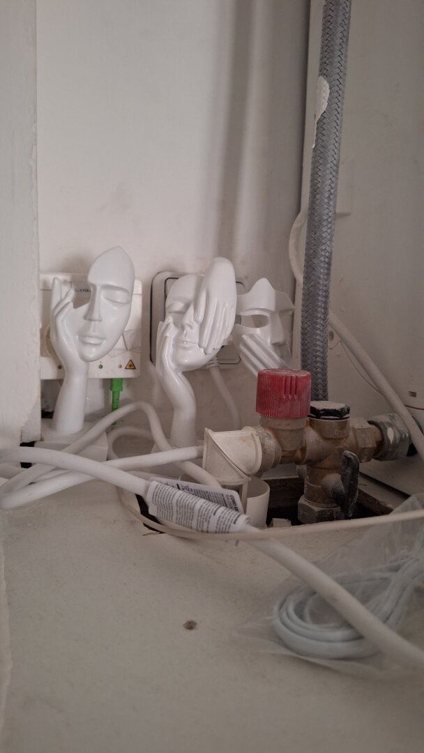 Jumble of electrical cables and plugs. Three face-like masks, with hands on them as if to convey worry, stuck on wall as if commenting on state of affairs