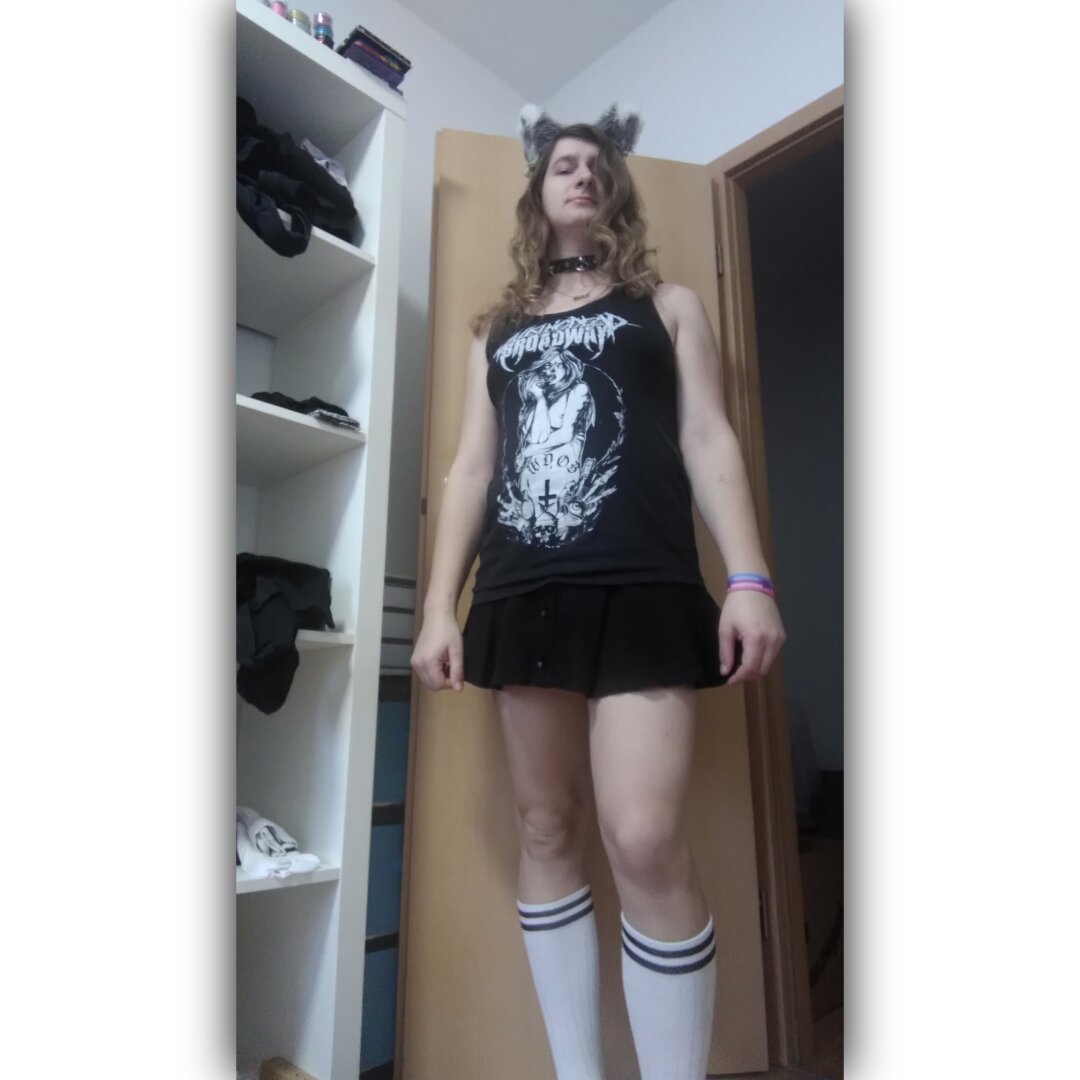 Photo of me, a white person with small breasts, standing in a white room with an open door and a white shelf with several clothes in the background.
I'm wearing long, brown, curly hair, grey cat ears with white tips, white ribbons and silver bells, a black collar with rivets and a ring for a leash, a black tank top, a black mini skirt and white tennis socks. On the tank top there is a white print of a nude person with breasts having the letters WDOB and a turned cross tattooed on their stomach and the band logo of Walking Dead on Broadway is over the person. I wear a catgender pirde wristband on my left wrist.
The photo is taken from diagonally below. That makes me look arrogant.