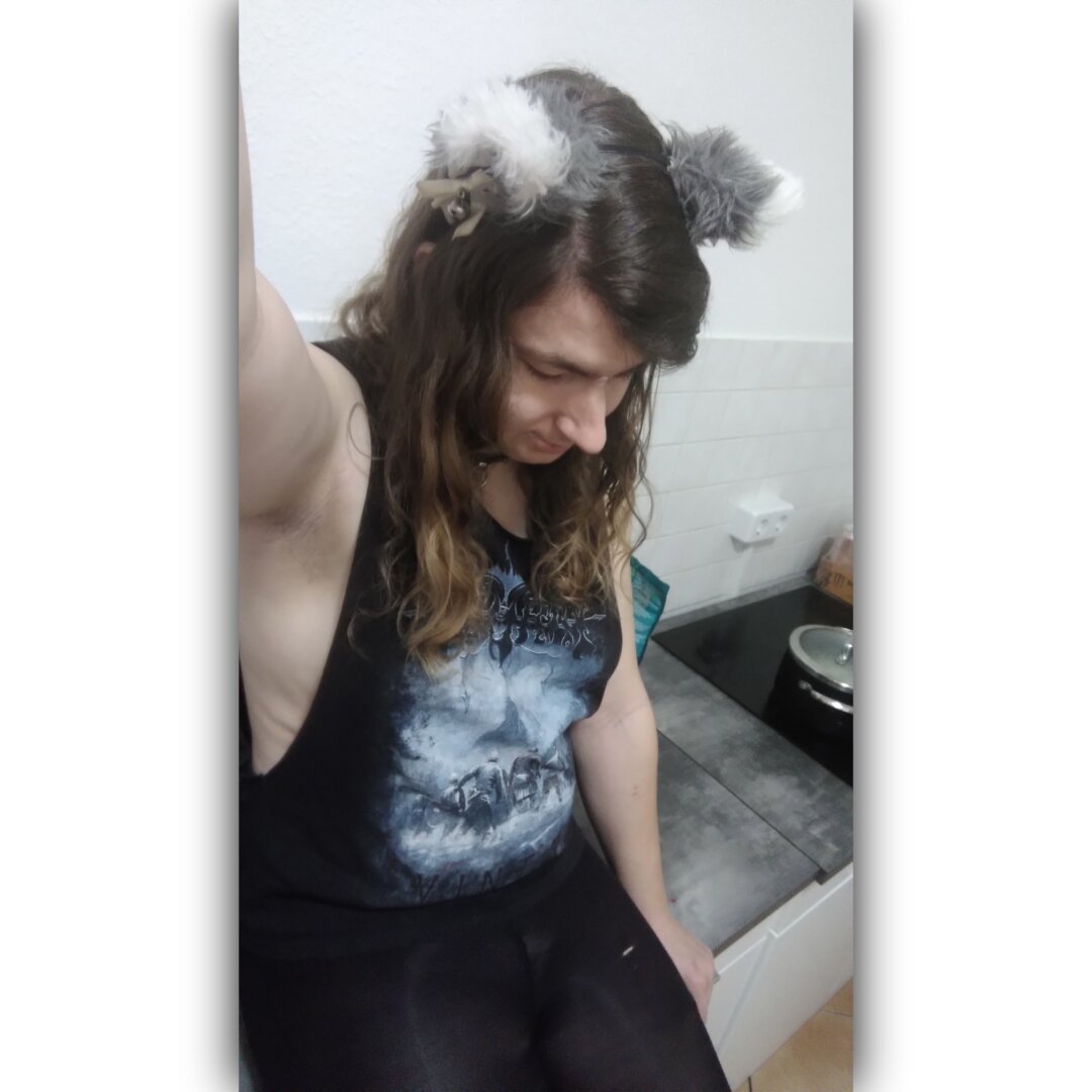 Selfie of me, a white person with small boobs, sitting on the kitchen furniture. I'm wearing long, brown, curly hair, grey cat ears with white tips, white ribbons and silver bells, a black top with wide cut outs for the arms, so you can see side boobs, and a black leggings.