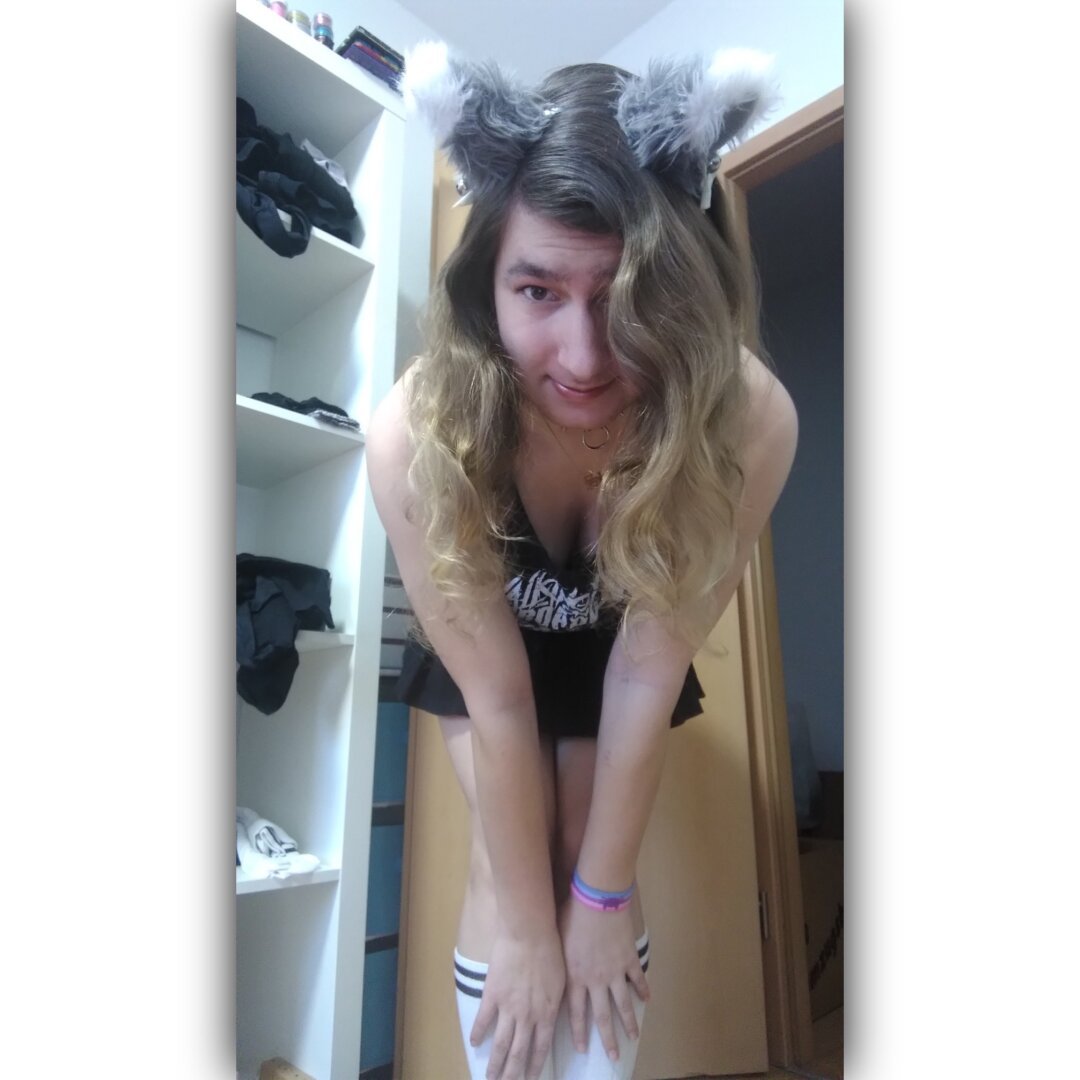 Photo of me, a white person with boobs, standing bent forward with my hands on my knees, looking in the camera.
I'm wearing long, brown, curly hair, grey cat ears with white tips, white ribbons and silver bells, a black tank top with a white print, which is nearly invisible, a black skirt, white tennis socks and a catgender pirde wristband on my left wrist.
In the background is a white shelf with several clothes and an open door.