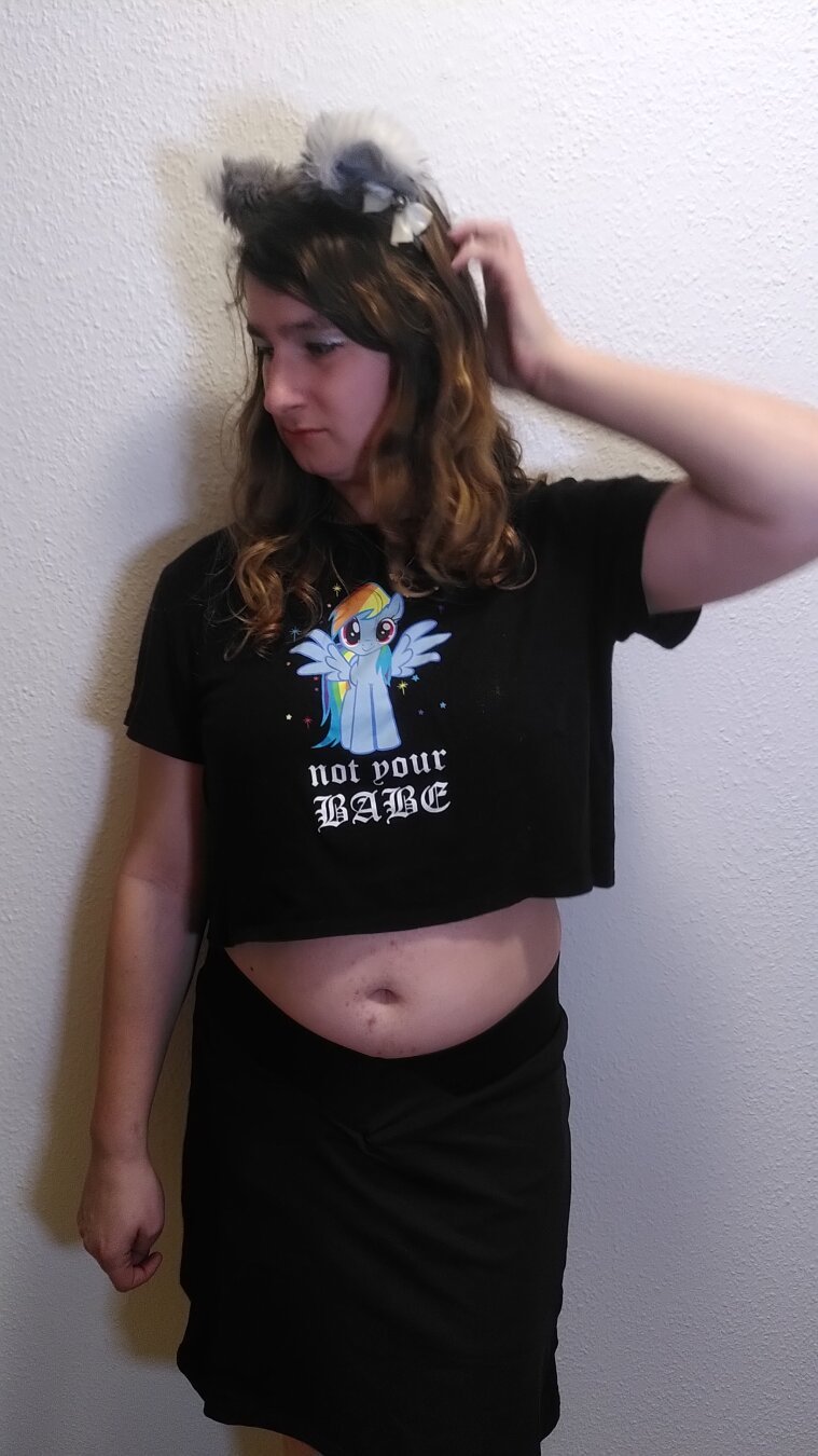 Photo of me, a white person with boobs, standing in front of a white wall. I'm wearing long, brown, curly hair, grey cat ears with white tips, white ribbons and silver bells, a black crop top and a black tennis skirt.
On the crop top is a print of Rainbow Dash from My Little Pony. She's a light blue pegasus pony with a rainbow colored mane. Under her is written in white letters 