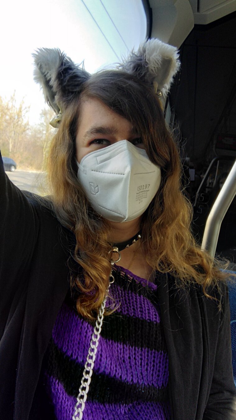 Selfie of me, a white person, in a tram.
I'm wearing long, brown, curly hair, grey cat ears with white tips, white ribbons and silver bells, a white FFP2 mask, a black collar with rivets and a leash on it and a black, purple striped knitted pullover and a black jacket that is open. I'm looking happy.