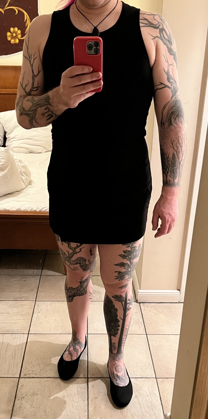 A person is taking a mirror selfie, wearing a black sleeveless shirt, a skort and black shoes. They have numerous intricate tattoos on their arms and legs, featuring tree and nature designs. The background includes a bed and some decorative elements on the wall.