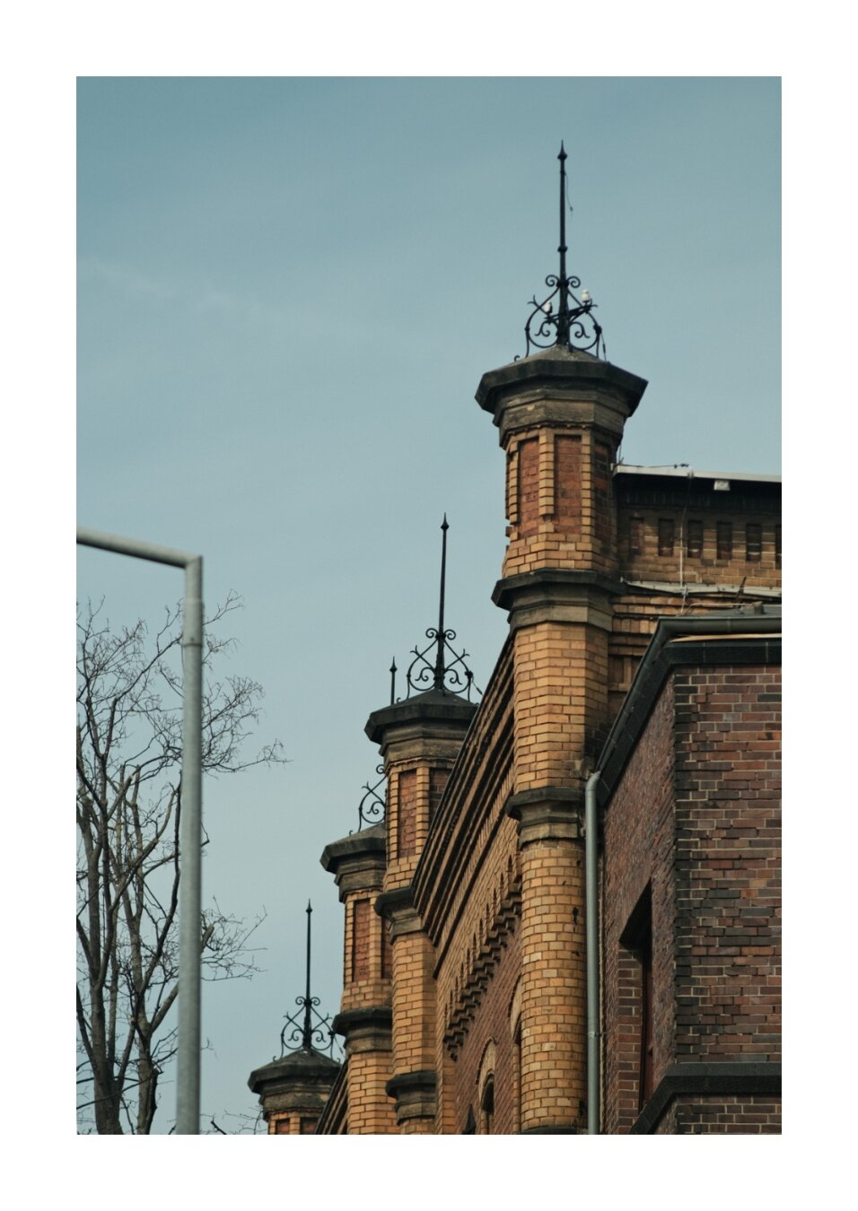 Detailansicht eines historischen Backsteingebäudes mit mehreren turmartigen Aufsätzen. Auf den kleinen Türmchen sitzen filigrane metallene Spitzen und ornamentale Verzierungen. Rechts ist die Fassade aus rotbraunem Backstein zu sehen, links ragen kahle Äste eines Baums und ein Teil eines Metallpfostens ins Bild. Der Himmel ist blassblau und wolkenlos.