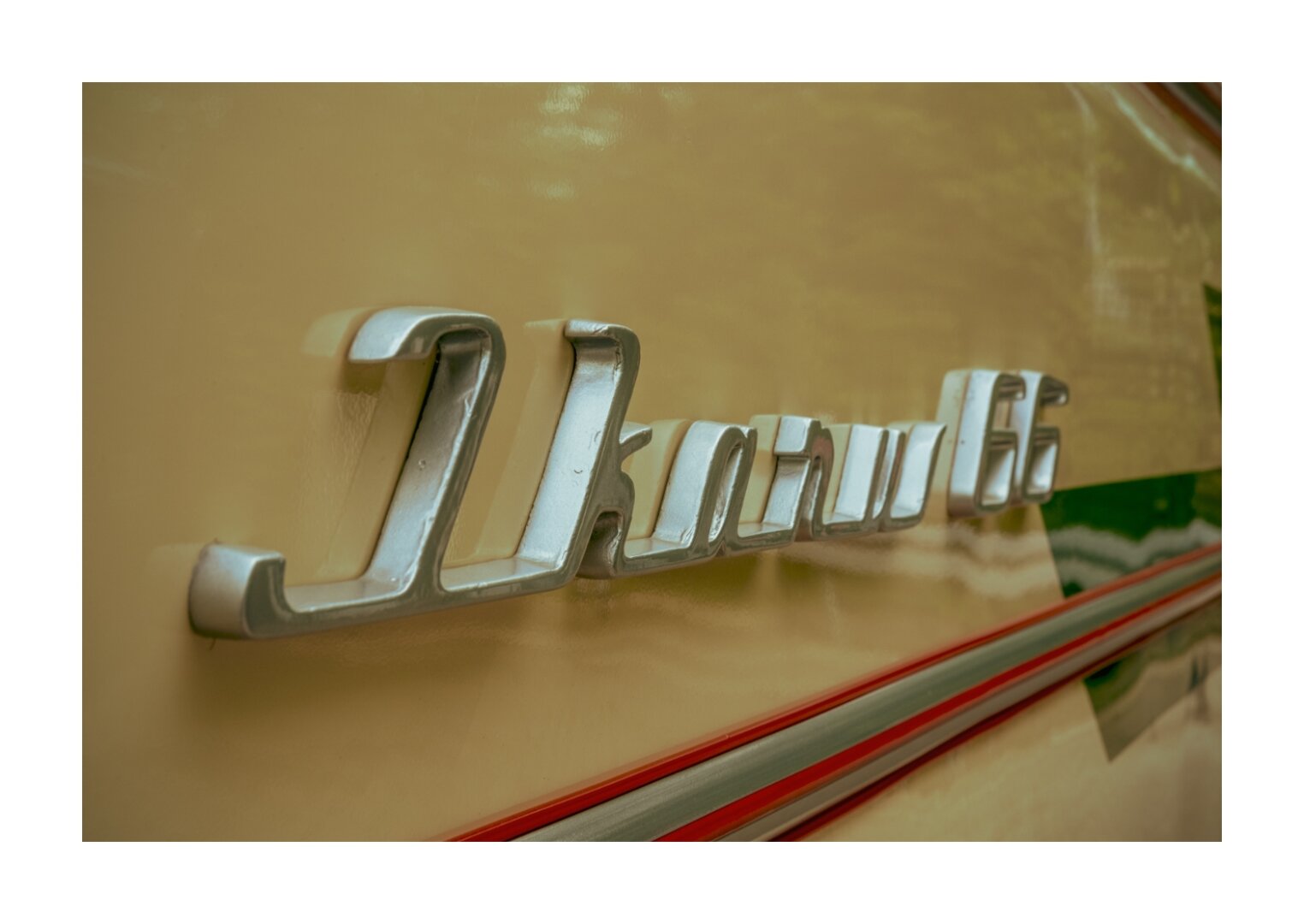 Nahaufnahme eines glänzenden, silbernen Schriftzugs "Ikarus 66" auf einer beigefarbenen Fahrzeugoberfläche. Der Schriftzug wirkt retro und ist in geschwungener, dreidimensionaler Typografie gehalten. Darunter verläuft ein roter Zierstreifen. Spiegelungen auf dem Lack deuten auf eine Umgebung im Freien hin.