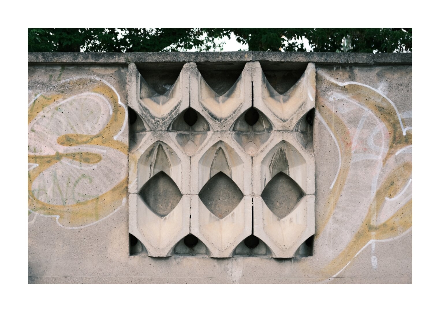 Betonwand mit einem eingelassenen, geometrisch geformten Sichtmuster aus wiederholten diamant- und bogenförmigen Elementen. Die Oberfläche ist verwittert und von hellgelbem und weißem Graffiti umgeben. Hinter den Öffnungen ist dunkler Hohlraum zu erkennen, darüber ragen Baumkronen in den Himmel.