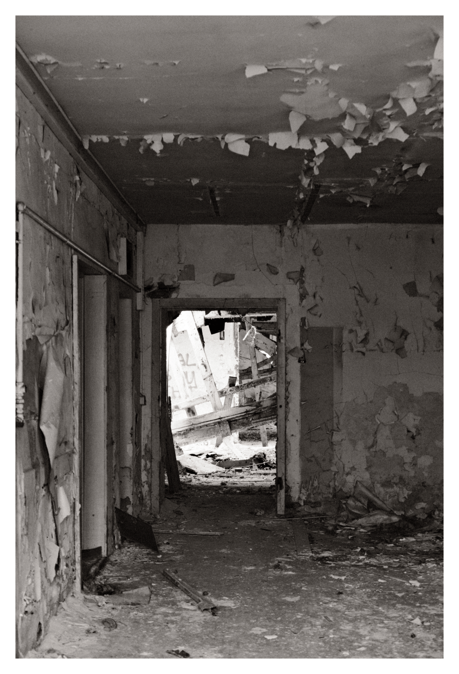 Innenraum eines stark beschädigten Gebäudes mit herabgefallener Decke, abblätternder Farbe und Trümmern auf dem Boden; Blick durch eine Tür in weiteren Verfall.