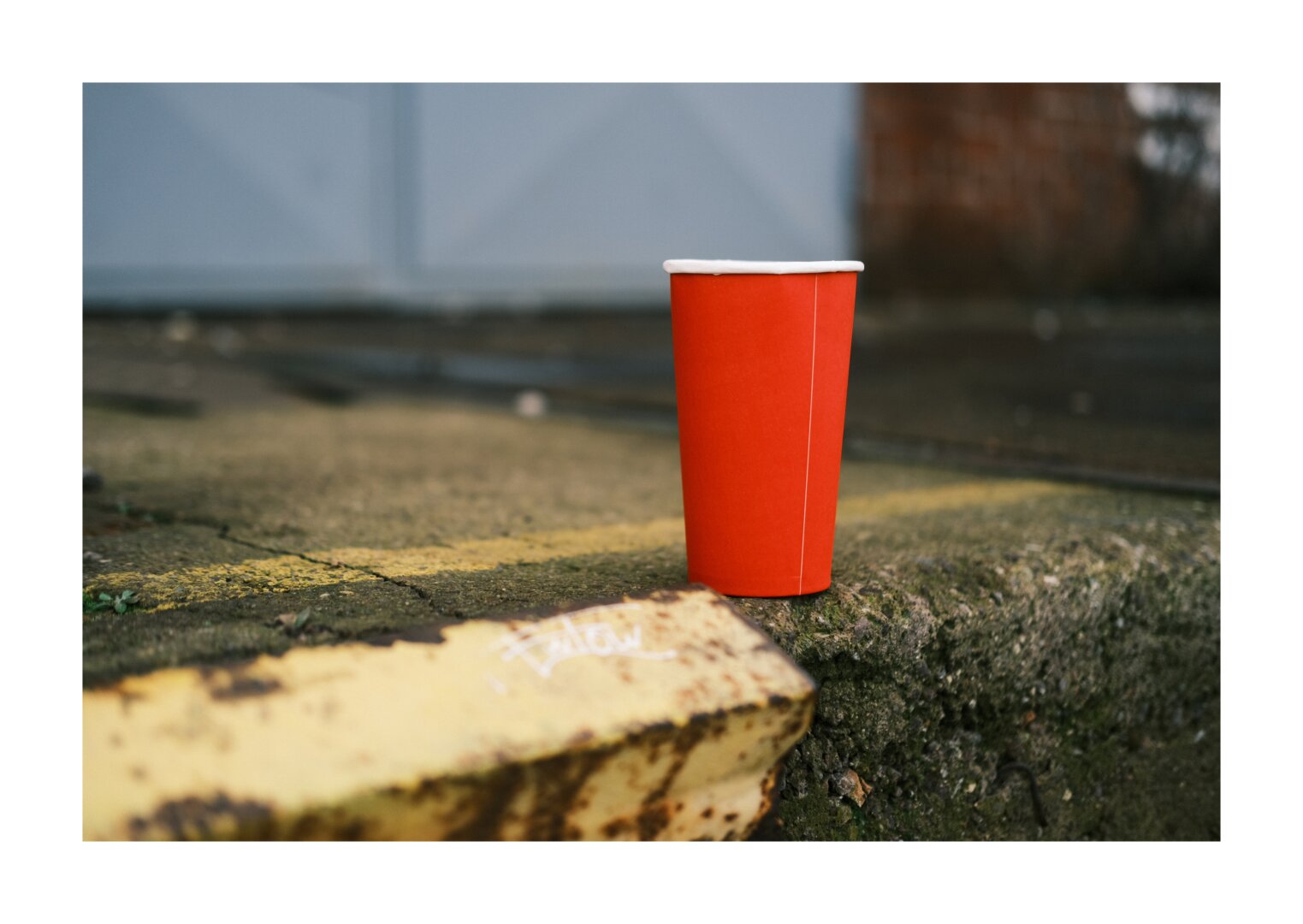 Ein roter Einwegbecher steht auf einer verwitterten Betonkante im Außenbereich. Der Boden ist uneben und mit Moos, Schmutz und einer verblassten gelben Markierung bedeckt. Im Vordergrund ist eine rostige Metallkante mit weißer Beschriftung zu sehen, der Hintergrund ist unscharf und zeigt ein großes graues Tor.