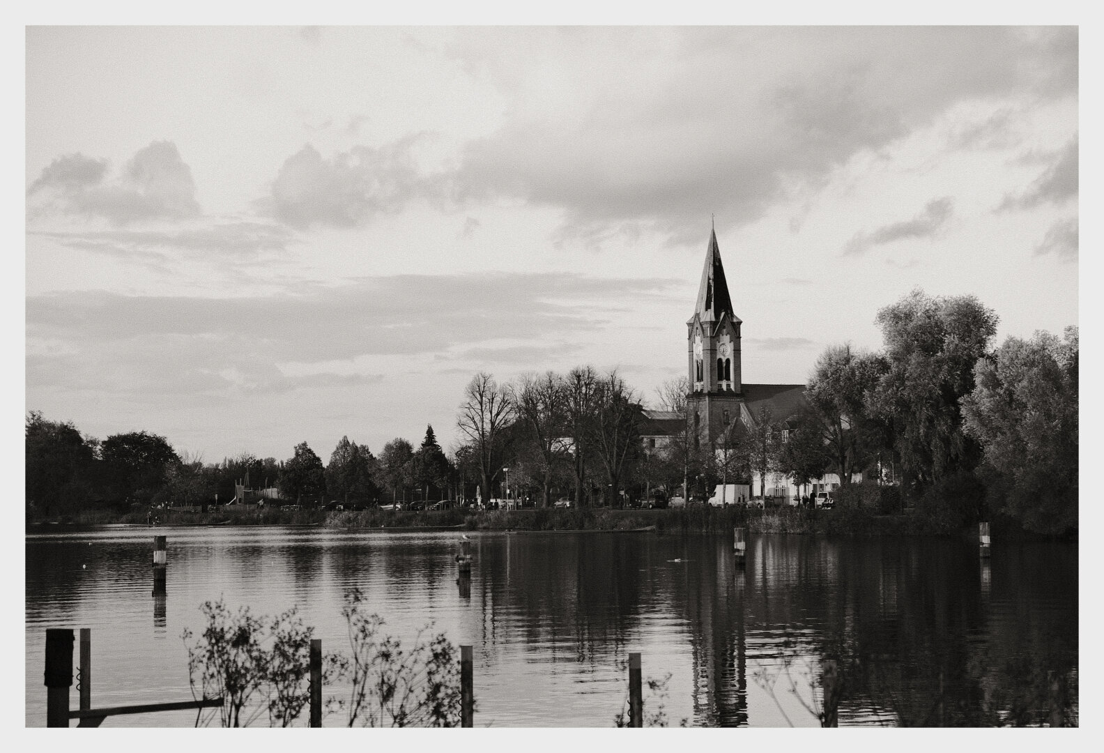 Schwarz-weiß-Landschaftsaufnahme eines ruhigen Sees, in dem sich Bäume und Pfosten spiegeln. Am gegenüberliegenden Ufer steht eine Kirche mit hohem, spitzem Kirchturm, umgeben von herbstlich kahlen und dichten Bäumen. Über dem Horizont hängen wolkige, leicht dramatische Himmelstrukturen.