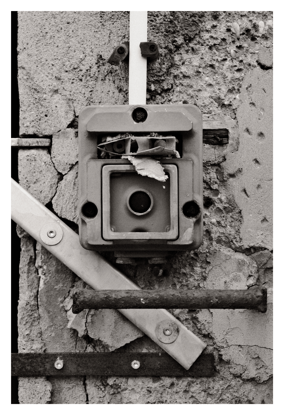 Nahaufnahme eines beschädigten Elektroschalters an rissiger Betonwand; Gehäuse gebrochen, Drähte und Papierfetzen sichtbar.