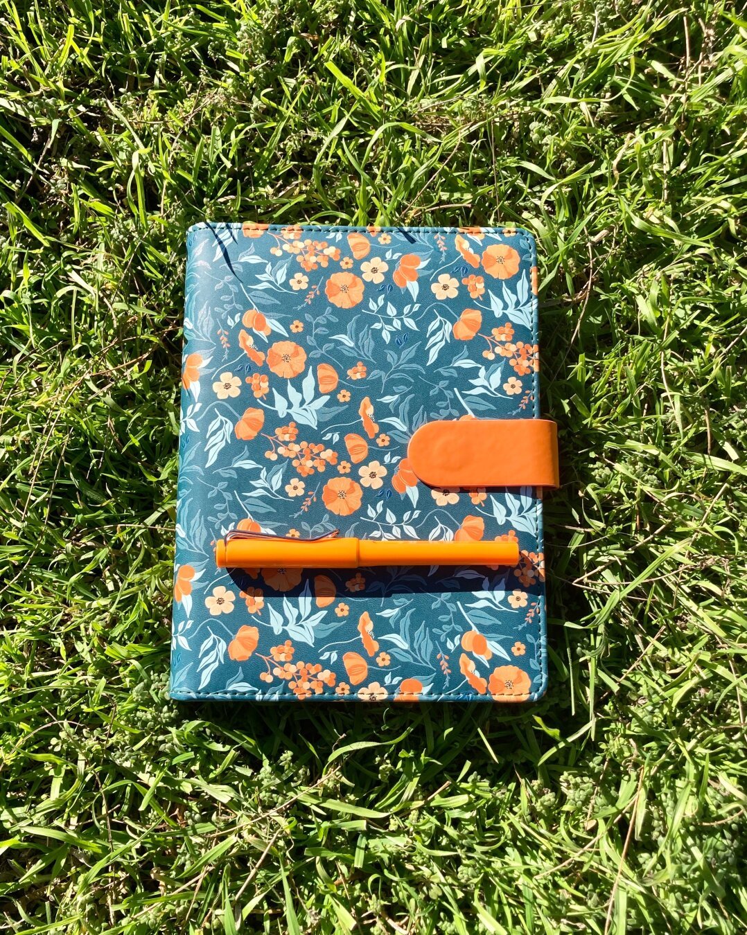 An orange fountain pen lying atop a green binder with an orange flower pattern, which is sitting on lush grass in bright sunshine