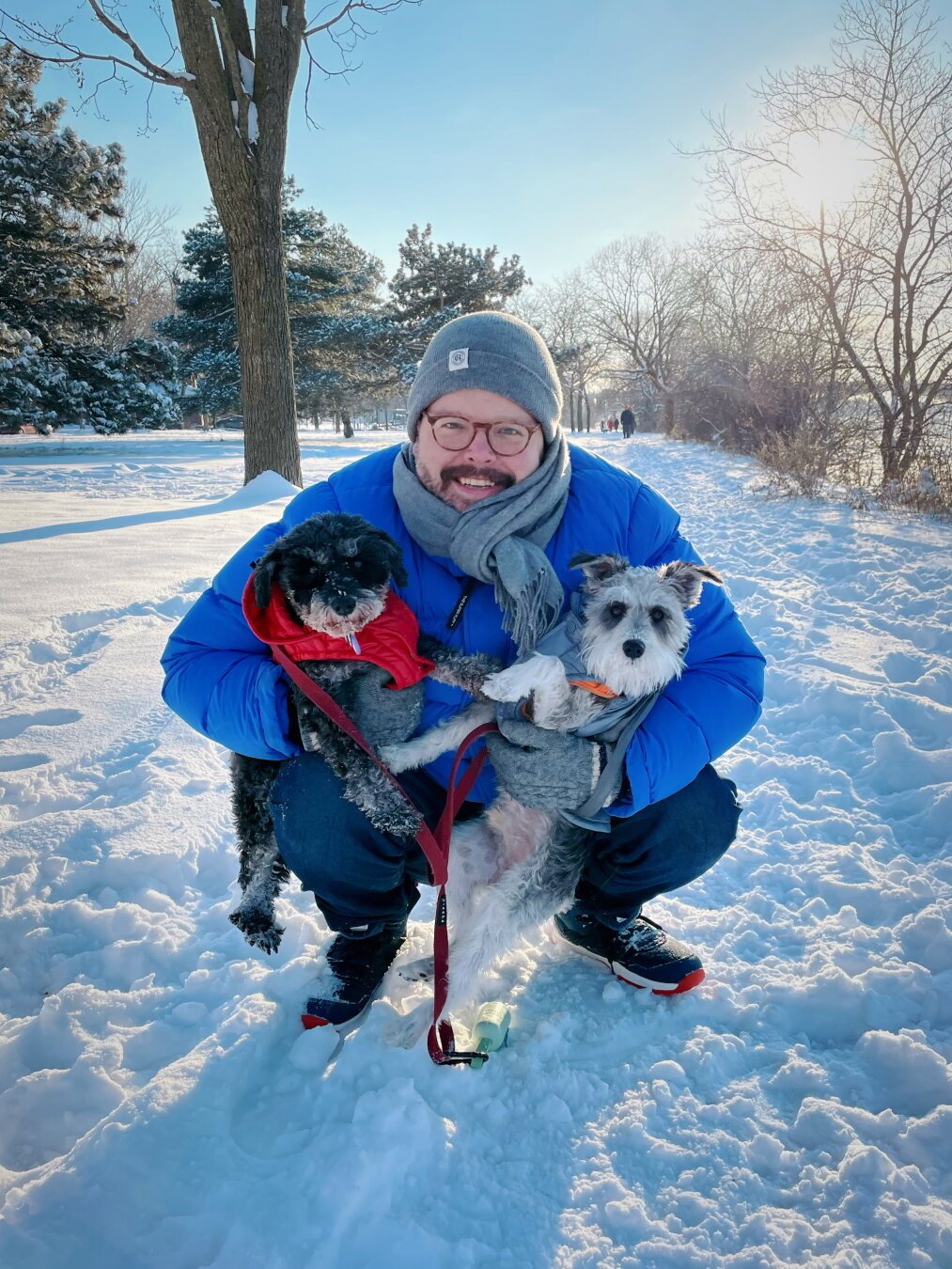Sunny winter day in a park, the ground is full of snow. White guy in a blue parka is squatting down and holding a miniature schnauzer in each arm. On the left, a black and silver with a red jacket, on the right, a salt and pepper with a gray jacket.