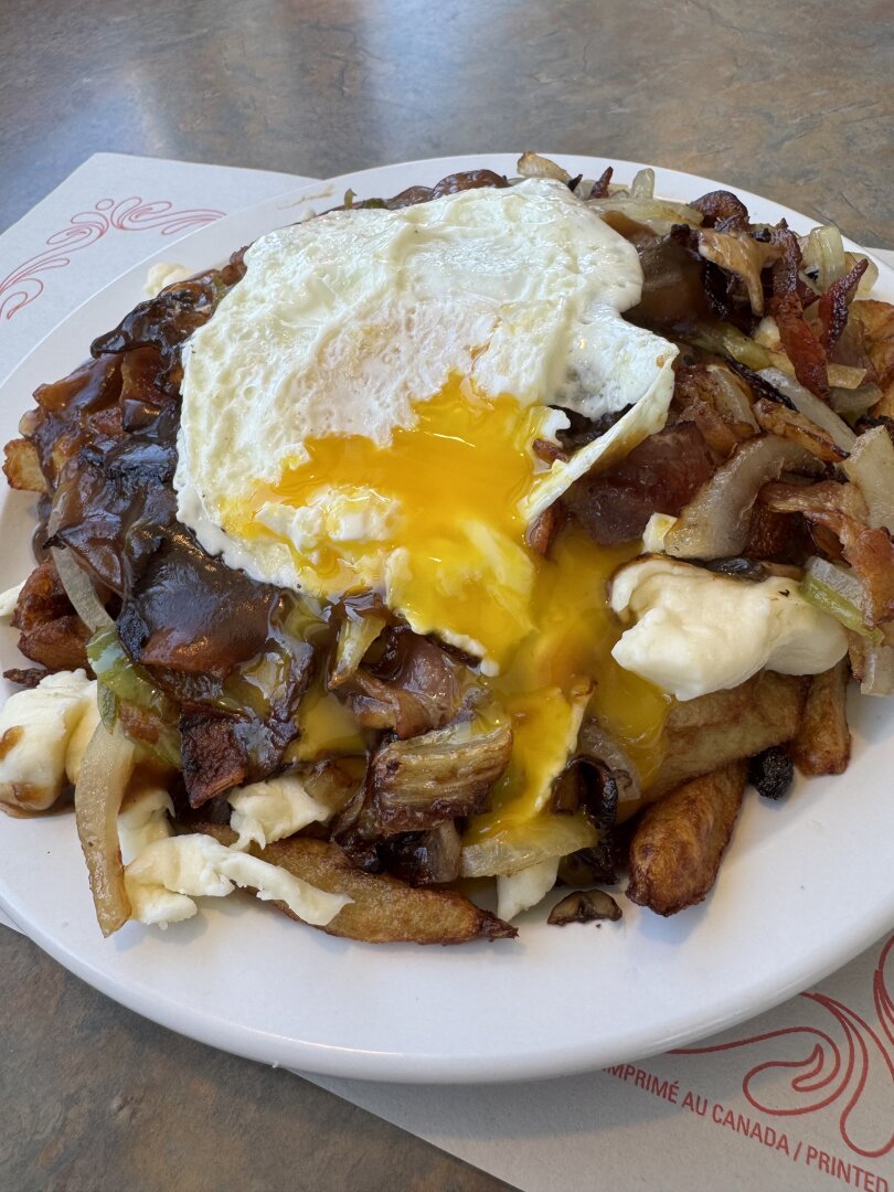 A poutine in a round white plate. There’s green peppers, mushroom, onion and bacon, with a runny over-easy egg on top.
