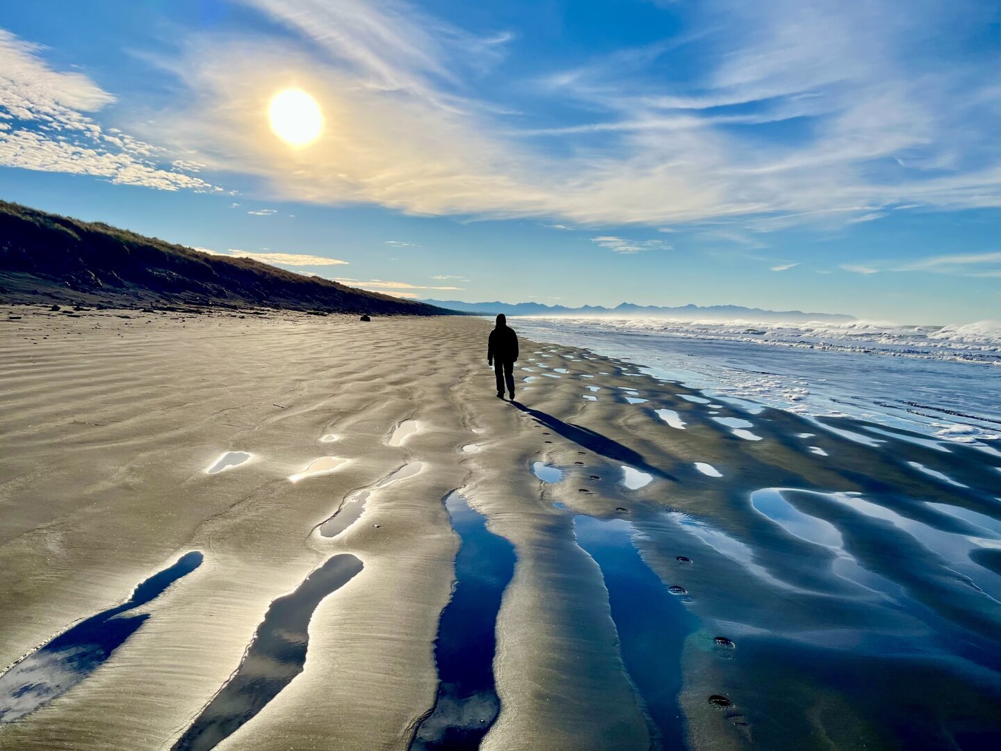 A hiker walks an ocean beach past wet ripples in the beach sand, toward a cloud-muted sun.