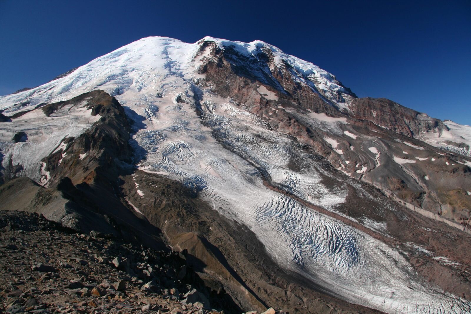 A large mountain of rocky ridges and ice under late summer sunshine and a blue sky. A long dirty white glacier curls down from the top.