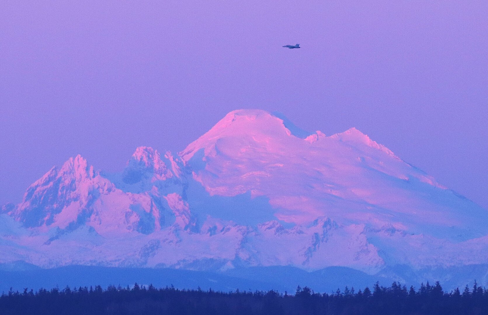 An EA-18 jet from Naval Air Station Whidbey Island flies in front of the volcano known as Kulshan, or Mount Baker, in the pinkish fading light of the day. The mountain is large, white with snow and conical. Two associated peaks are to the left, the Black Buttes, at this time mostly white with snow.