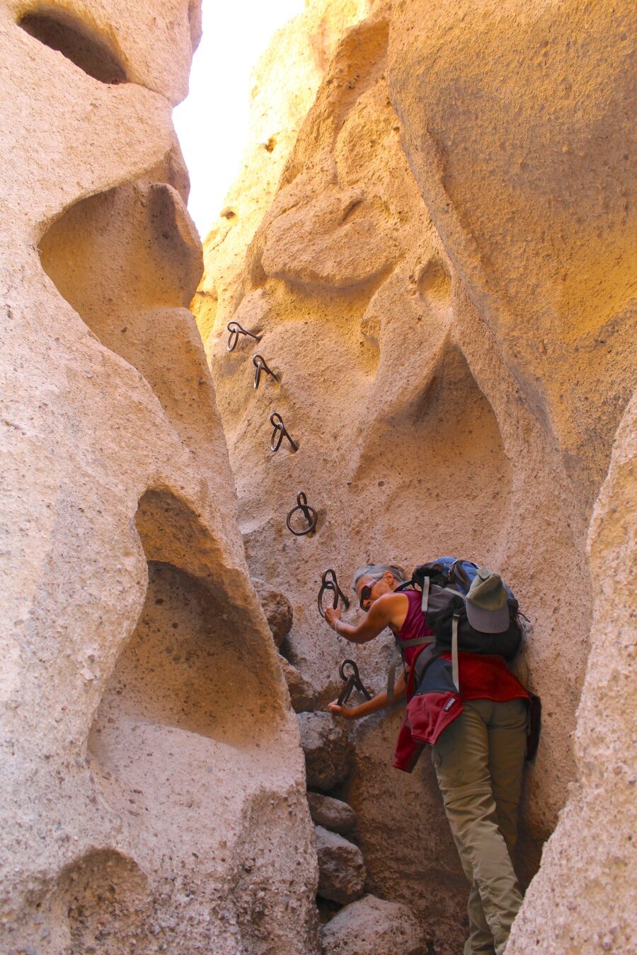 Hiker with her hair up wearing a small backpack and sunglasses looks back while beggining a climb up a rock slot canyon on a desert trail, with several steel rings imbedded in the rock as hand holds to assist hikers. The rock is a pale pink in color and there is sunlight glowing atop the slot about 20 feet up.