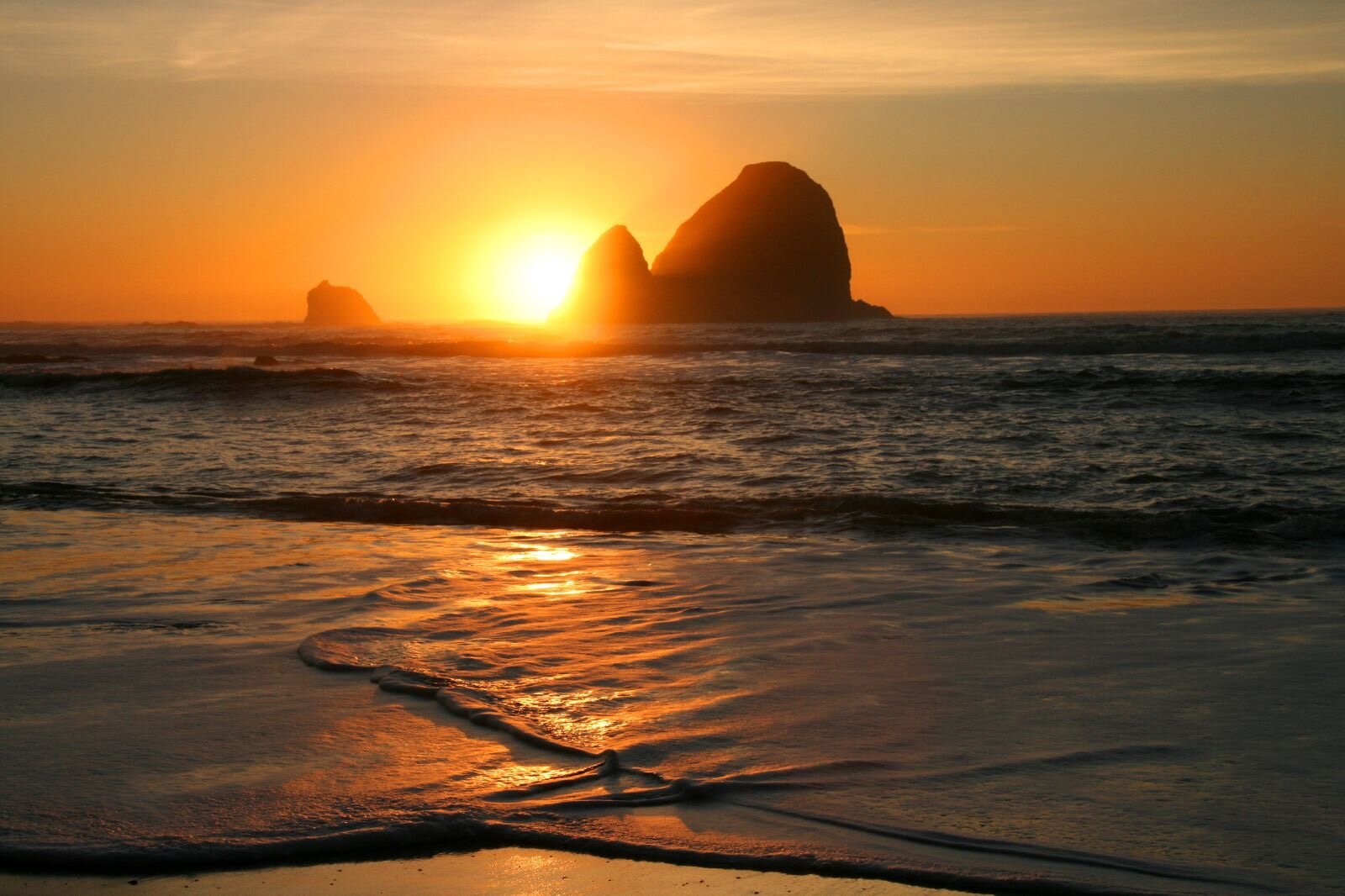 The horizon glows golden as the sun descends as a ball of fire beyond sea stacks in the ocean, its rays reflected in the shallows of a wave along shore.