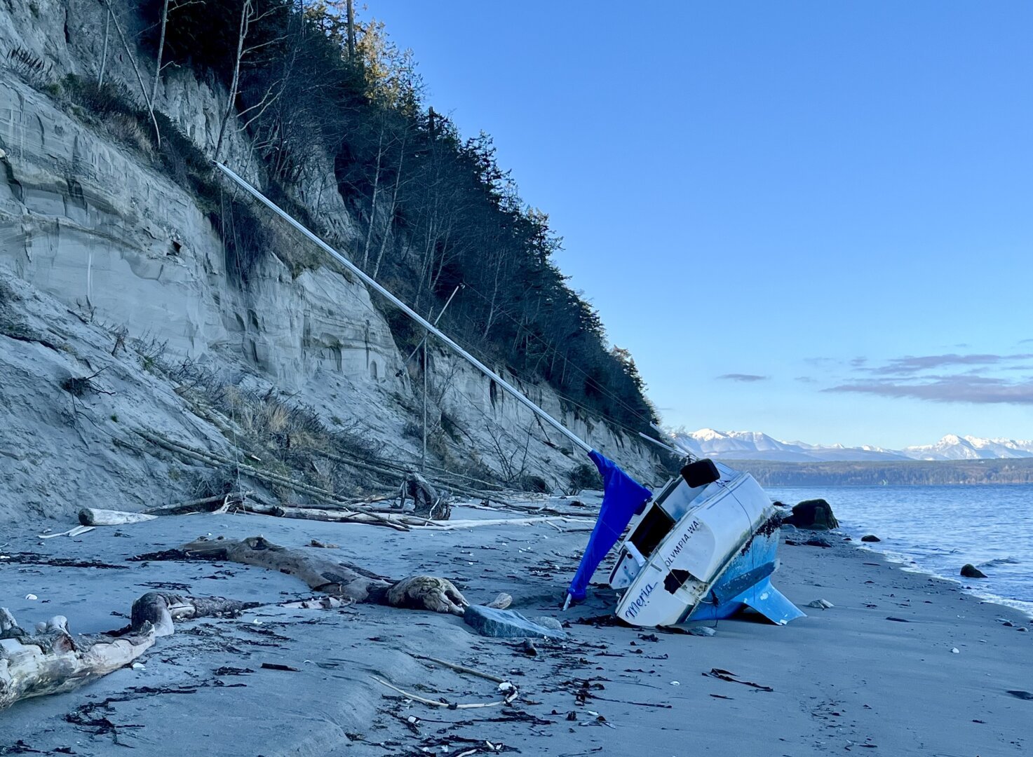 Photo looking at the stern end of a sailboat washed ashore and laying in her side, her mast at a 45 degree angle, on a beach below a steep, forested bluff. Water and blue sky on the right. On the back is her name, “Meria.”