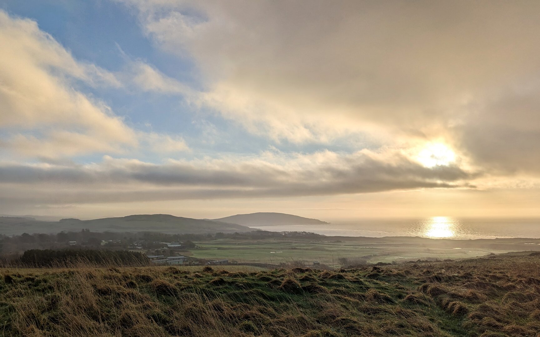 View from a hill over the coastline with a distant headland in silhouette and a dramatic morning sky. sun reflected in the sea.