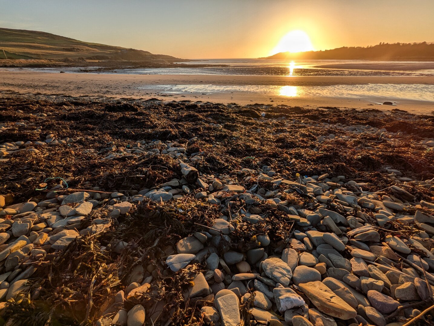 View of a beach looking out to sea, with sun setting over headland in background and stones and seaweed glowing in the warm light in the foreground