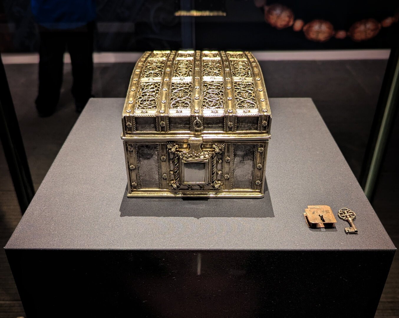 An ornate silver box with a lock and key beside it in a display case with museum lighting showing the details of the decoration. The box is a bit bigger than A4 in footprint and about four inches high.