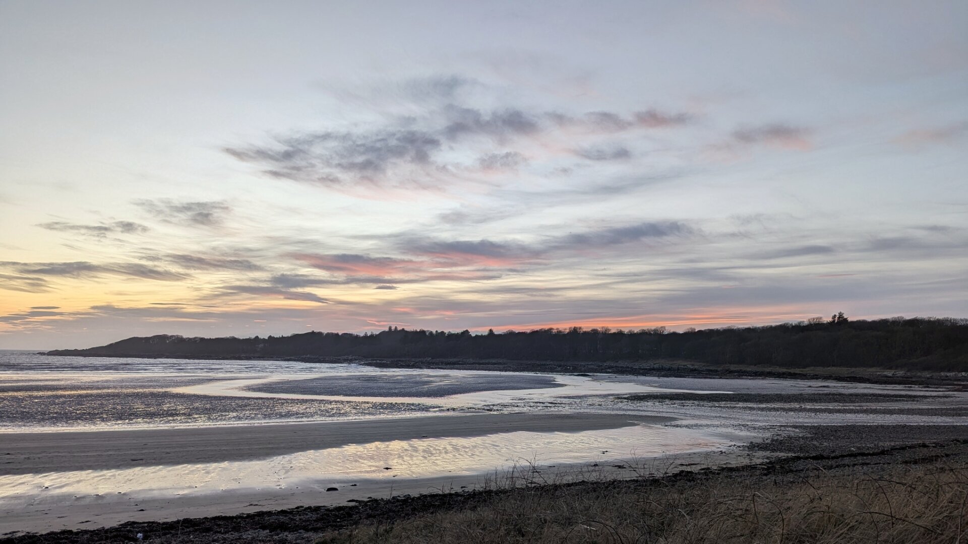 Clouds tinged with pink and yellow above a headland in silhouette looking a little like a sleeping dragon. Foreground a beach at low tide with sand banks and shallow water reflecting sky.