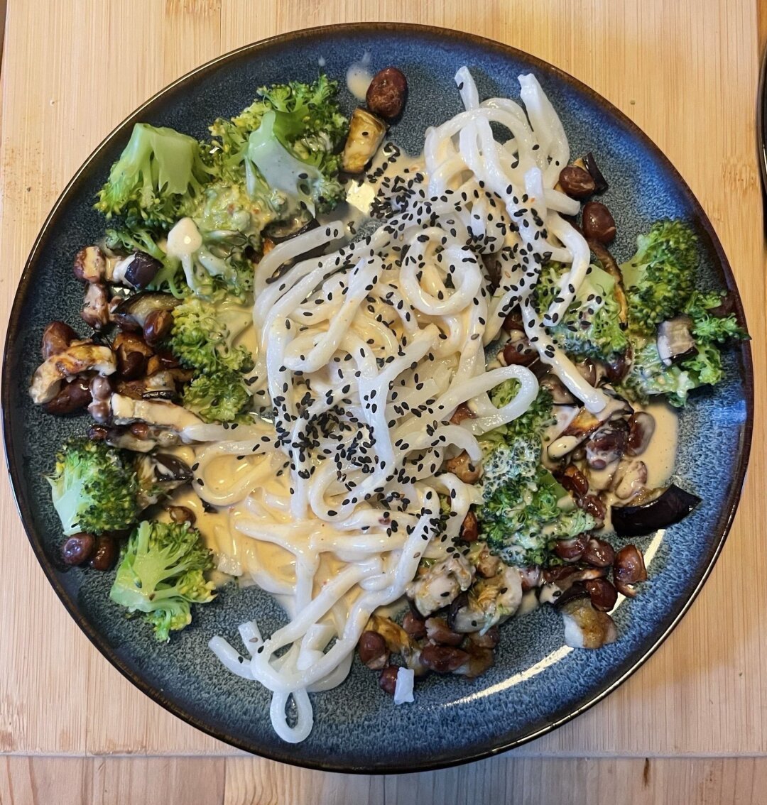 Ein runder blauer Teller mit Nudeln mit schwarzem Sesam, umgeben von gedämpftem Brokkoli und gemischtem sautiertem Gemüse und Bohnen. Die Schale wird auf einer hellen Holzoberfläche präsentiert.
