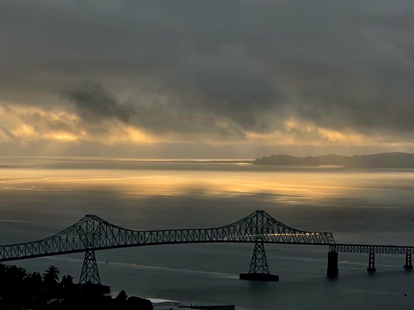 Bridge over a river in the foreground, dimly lit, water beneath dark, with the sun breaking through the clouds in the distance, shining brightly on the water.