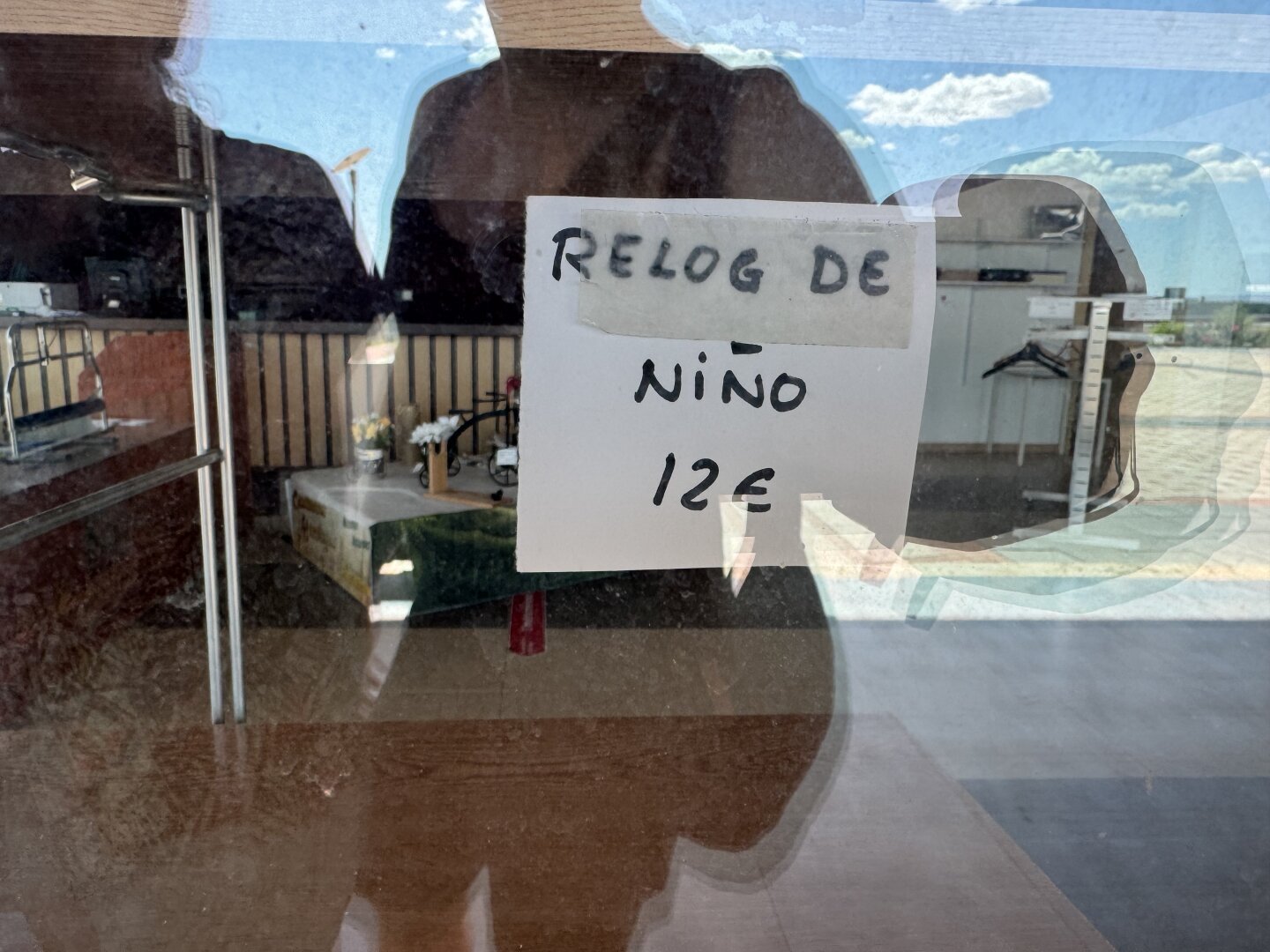 Reflejo de escaparate con un post-it pegado diciendo “Relog (escrito con G) de niño 12 euros”