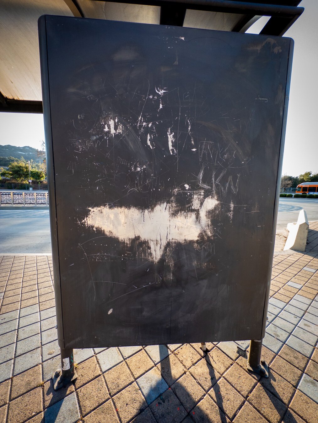 The back of a sign at a bus stop. It’s painted brown, with a large scrape that alllmossst looks like a Batman logo.