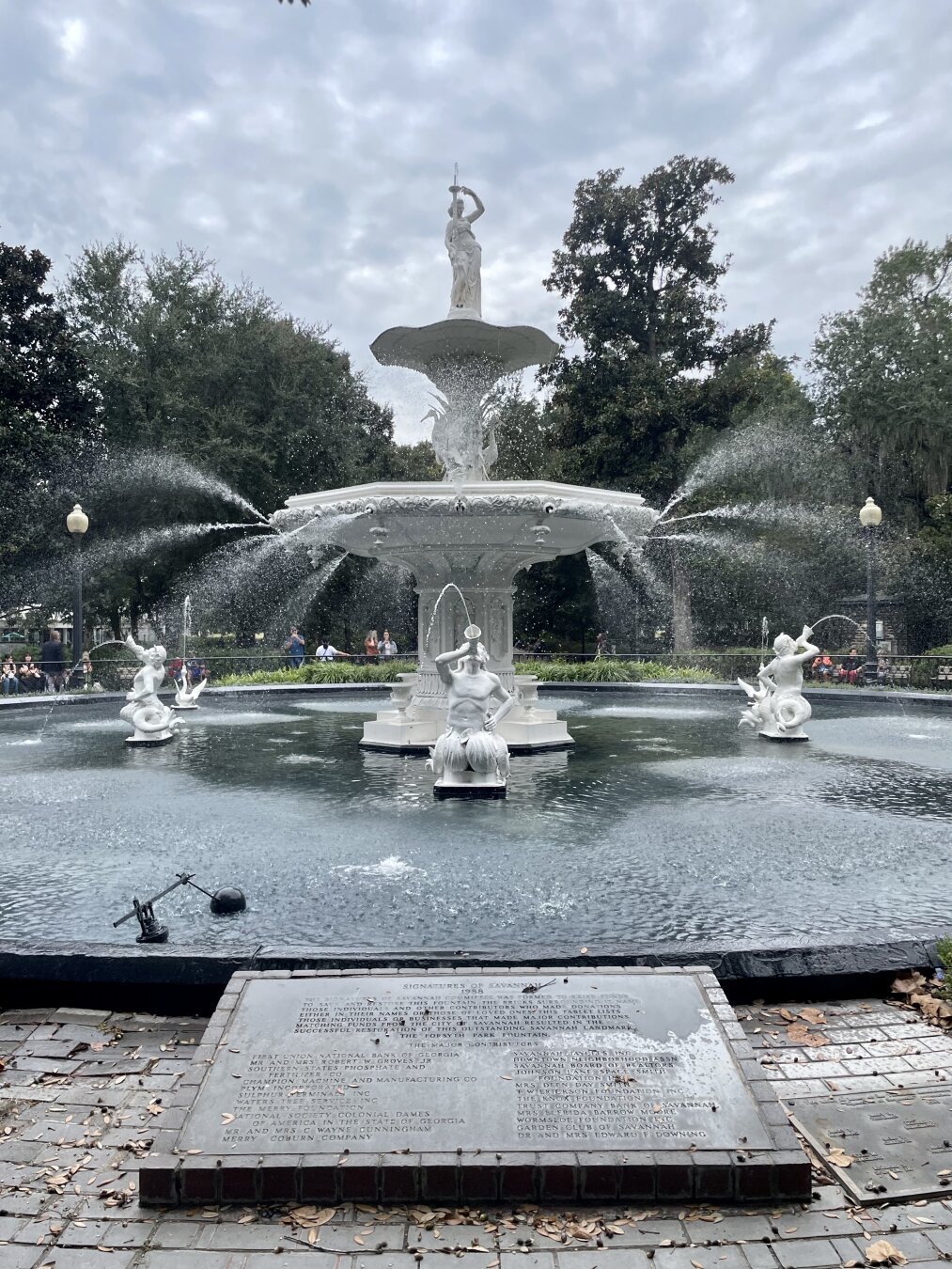 A view of the fountain in Forsyth Park in Savannah. The white fountain is turned on and jets of water stream outward and into the pool that encircles the fountain. Merfolk blow water from their trumpets, and the figure of a woman stands atop the fountain with two birds that look like herons decorating the spot just below her.