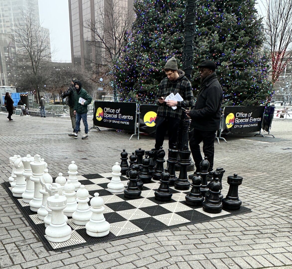 A large chess set with movable pieces sits on the sidewalk of a downtown area while two men exchange information in the background. The city Christmas tree stands behind them on a foggy day.