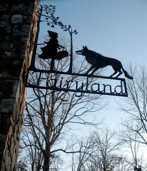 A metal sign depicting a cut out of little red riding hood, being confronted by a wolf. Underneath, the lettering spells Fairyland in script. It is attached to a stone fence pillar.