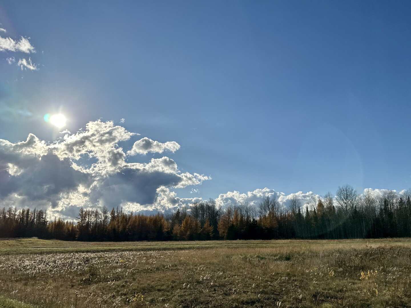 The sun shines onto white clouds in a blue sky over autumn fields. Trees are visible in the distance.