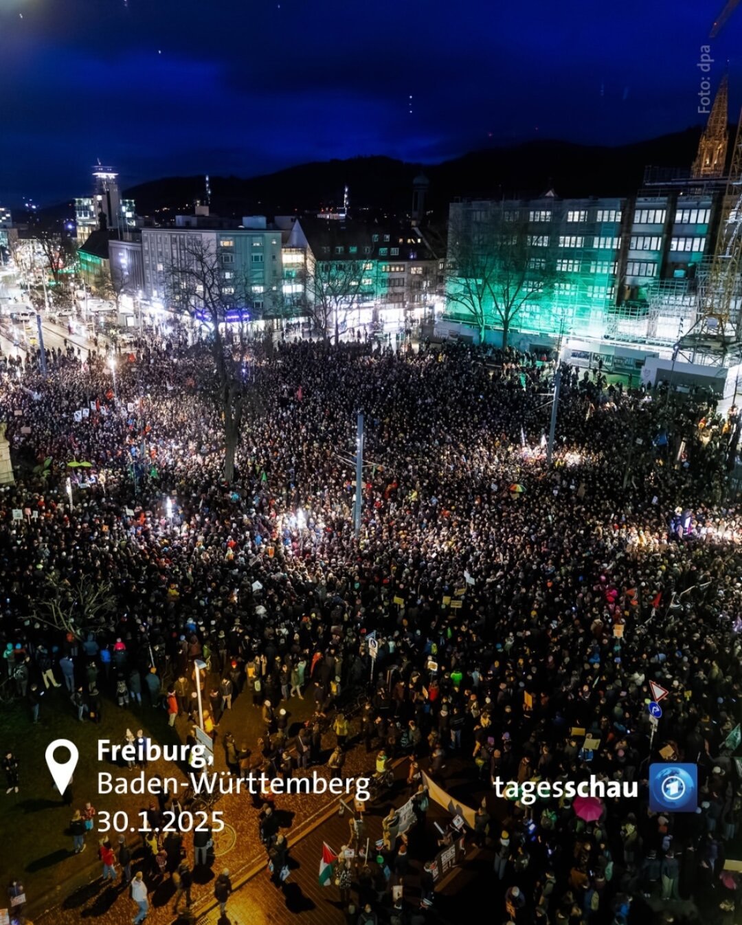 Eine riesige Demonstration in der Innenstadt von Freiburg, Baden-Württemberg, am 30. Januar 2025. Es wurde am Abend aufgenommen, der Himmel ist dunkelblau, und die Stadtbeleuchtung erhellt die Straßen. Tausende von Menschen haben sich auf einem großen Platz versammelt, der von modernen Gebäuden mit beleuchteten Fenstern umgeben ist.

Die Menge ist dicht gedrängt, viele Teilnehmende halten Schilder und Banner hoch, die jedoch aus der Distanz nicht lesbar sind. Einige Bereiche der Demonstration sind besonders hell erleuchtet, möglicherweise durch Scheinwerfer oder Kameras. Die Straßen und Gebäude in der Umgebung sind ebenfalls gut beleuchtet, einige Fassaden sind grün angestrahlt.

In der unteren linken Ecke des Bildes befinden sich weiße Texteinblendungen mit Informationen zur Stadt (Freiburg, Baden-Württemberg) und zum Datum (30.1.2025). In der unteren rechten Ecke befindet sich das Logo der „Tagesschau“, was darauf hinweist, dass das Bild von einem Nachrichtenmedium stammt.