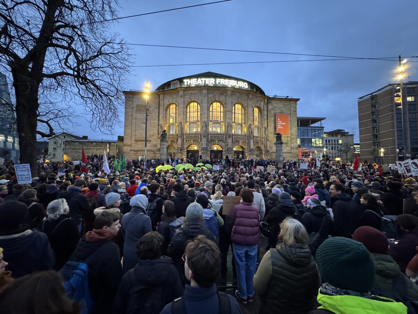 Eine große Demonstration vor dem Theater Freiburg in der deutschen Stadt Freiburg. Es ist Abend oder frühe Dämmerung, und das Gebäude des Theaters ist hell erleuchtet, während der Himmel bereits dunkelblau ist.

Eine große Menschenmenge versammelt sich auf dem Platz vor dem Theater. Viele der Demonstrierenden tragen Winterkleidung wie Mützen, Schals und dicke Jacken. Zahlreiche Menschen halten Schilder und Banner hoch, auf denen teilweise Schriftzüge zu erkennen sind, obwohl die genauen Inhalte nicht vollständig lesbar sind. Es sind verschiedene Fahnen zu sehen, darunter rote und grüne Fahnen. Einige Menschen in der Mitte der Menge halten auffällige gelbe Regenschirme.

Im Hintergrund sind moderne Gebäude und Straßenlaternen zu erkennen, die die Szene beleuchten. Die Atmosphäre wirkt friedlich, aber auch entschlossen, da die Menge konzentriert scheint und aufmerksam nach vorne schaut, möglicherweise auf eine Rede oder eine Aktion auf einer nicht sichtbaren Bühne.