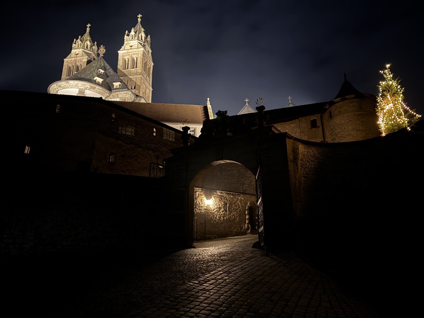 Comburg bei Nacht, mit einem großen Torbogen im Vordergrund, durch den ein Kopfsteinpflasterweg führt. Die Szenerie ist stimmungsvoll beleuchtet, mit einer warmen Lampe am Tor und einem geschmückten, leuchtenden Weihnachtsbaum rechts. Im Hintergrund erheben sich zwei imposante Kirchtürme mit Kreuzspitzen, die von unten angestrahlt werden und sich gegen den dunklen Nachthimmel abheben.