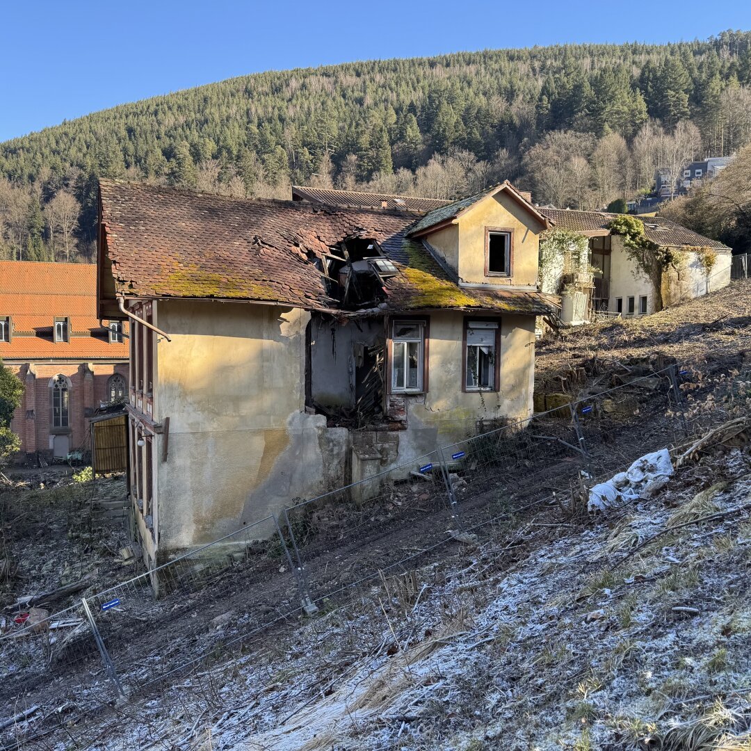 Ein altes, verlassenes Haus an einem Hang. Das Gebäude ist stark beschädigt: Das Dach hat ein großes Loch mit eingestürzten Teilen, und die Wände sind verwittert und teilweise eingerissen. Moos und Pflanzen wachsen auf dem Dach. Die Fenster sind teilweise kaputt oder fehlen ganz.

Im Vordergrund ist der Hang leicht mit Frost oder Raureif bedeckt, und ein provisorischer Bauzaun steht schief um das Gelände. Im Hintergrund sieht man eine bewaldete Hügelkette sowie ein größeres Gebäude mit einem roten Dach, das wie eine Kirche oder ein historisches Bauwerk aussieht. Die Sonne scheint, und der Himmel ist klar blau.