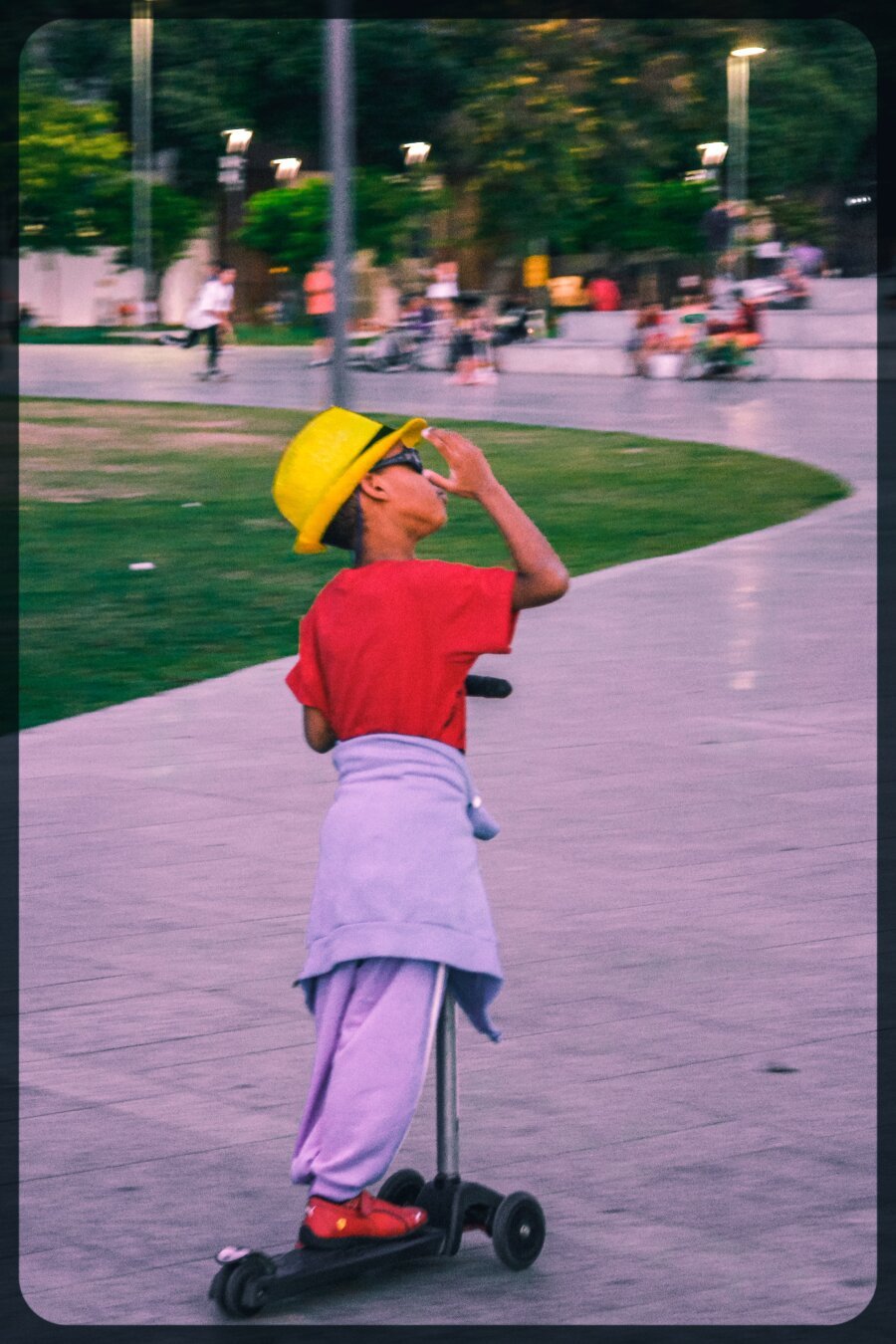 A very stylish boy skating around. His hat is yellow and he wears dark glasses and red shirt.
