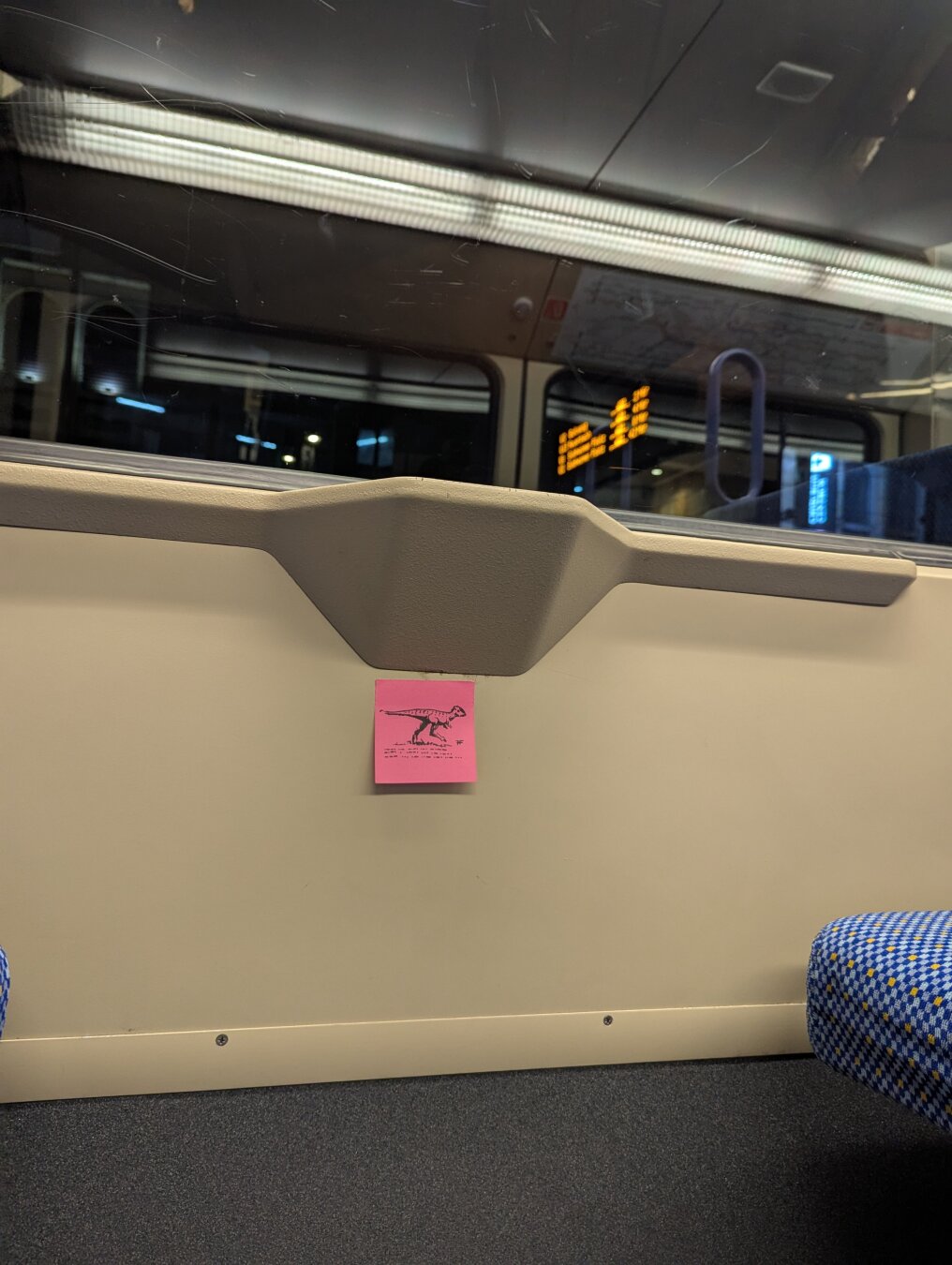 A dinosaur drawn onto a post-It note beneath the window inside a Stuttgart stadtbahn.