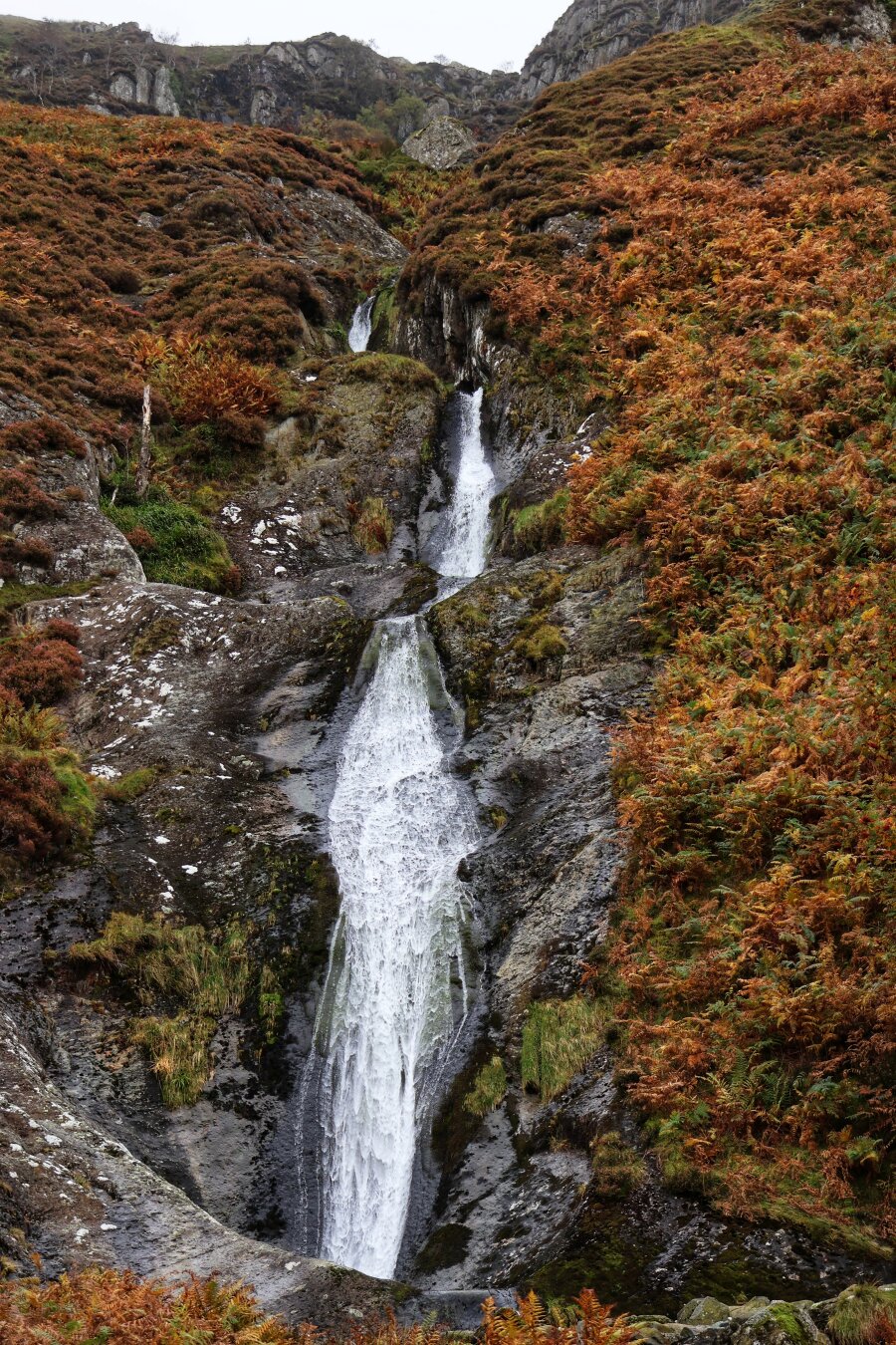 A breathtaking view of a slender waterfall cascading down a rocky gorge, surrounded by rugged, moss-covered cliffs. The waterfall splits into multiple streams as it descends, creating a delicate, misty effect against the dark, damp rock face. The surrounding landscape is adorned with vibrant autumnal foliage, featuring rich hues of orange, red, and green vegetation. The scene exudes a sense of tranquillity and the raw beauty of nature in a mountainous or upland setting.
