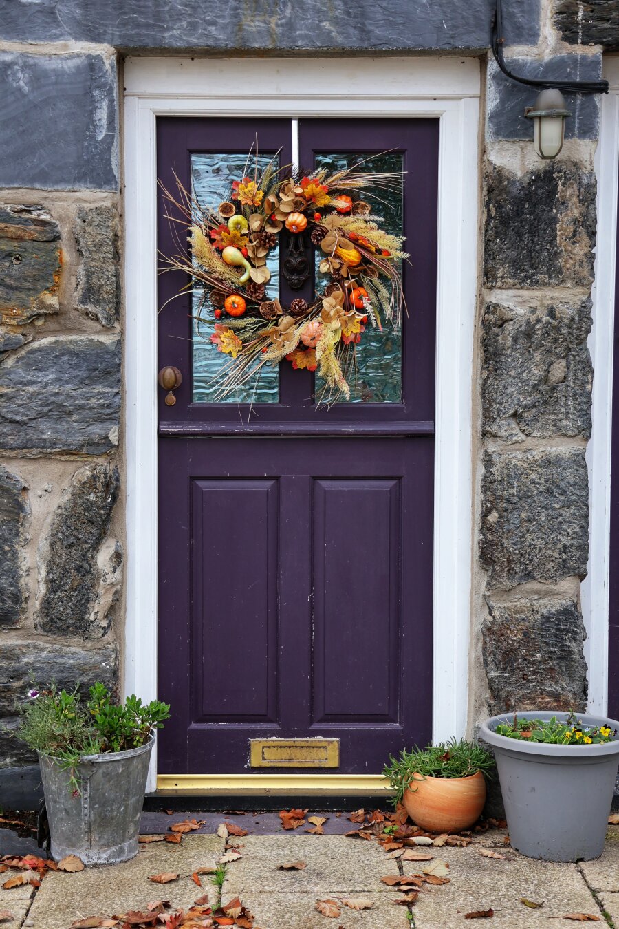 A charming, deep purple half-glazed door set into a rustic stone wall, adorned with an autumnal wreath featuring dried wheat, miniature pumpkins, pinecones, and colourful leaves. The door is framed in white and flanked by two potted plants—one with green foliage and the other with blooming yellow flowers. Scattered autumn leaves rest on the stone step below, enhancing the seasonal ambiance.